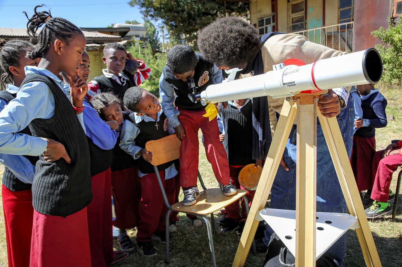 A group of Ethiopian schoolchildren in uniform watch with enthusiasm as a classmate, standing on a chair, looks through an astronomical telescope mounted on a wooden tripod. A curly-haired instructor guides the activity, ensuring that the students understand how to use the telescope.